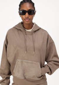 Sudadera marrón de tejido suave con un acabado texturizado, que presenta un bolsillo frontal y detalles de costura dorada; el modelo lleva gafas de sol negras.