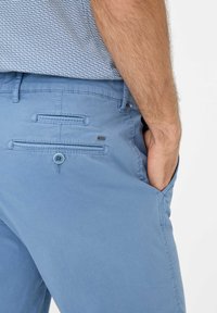 Chinos azzurri chiari in cotone con una superficie liscia. Dispone di due tasche posteriori, una delle quali con chiusura a bottone, e un dettaglio del logo del brand.