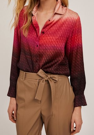 Femme portant une chemise à manches longues dégradée du rouge au violet foncé avec des pois subtils, rentrée dans un pantalon taille haute beige avec une ceinture nouée.