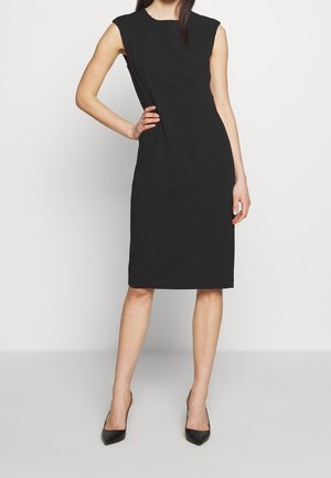 Vrouw draagt een mouwloze, knie-lange zwarte jurk en zwarte pumps met spitse neus, staande met één hand op haar heup tegen een effen achtergrond.