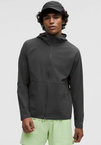 Donkergrijze waterdichte herenjas met capuchon, ritssluiting aan de voorkant en zakken. Draagover een lichtgroene korte broek, met een gladde, matte afwerking.