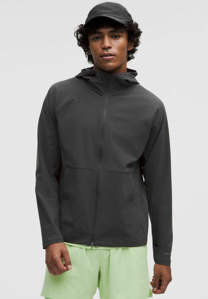 Donkergrijze waterdichte herenjas met capuchon, ritssluiting aan de voorkant en zakken. Draagover een lichtgroene korte broek, met een gladde, matte afwerking.