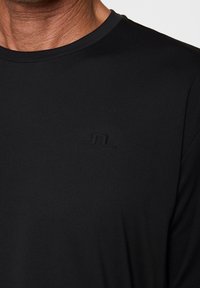 Närbild av en person som bär en svart rundhalsad T-shirt med en subtil pr präglad logotyp på bröstet.