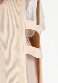 Un vêtement en tricot côtelé crème présente des côtés ouverts avec des sangles horizontales. Une blouse intérieure lisse en tissu léger est partiellement visible en dessous.