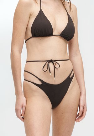 Femme portant un bikini string noir avec un haut triangulaire et un bas noué devant, montrant des bretelles latérales découpées sur un fond uni.
