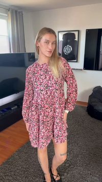 Mlada žena s dugom plavom kosom, odjevena u ružičastu haljinu s cvjetnim uzorkom i crne cipele, stoji u dnevnoj sobi s televizorom i umjetničkim djelima na zidu.