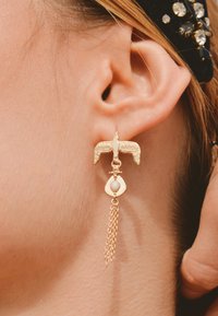 Boucles d'oreilles en or en forme d'oiseau avec un accent perlé, présentant une chaîne pendante et une goutte ouverte derrière la perle, finition texturée.