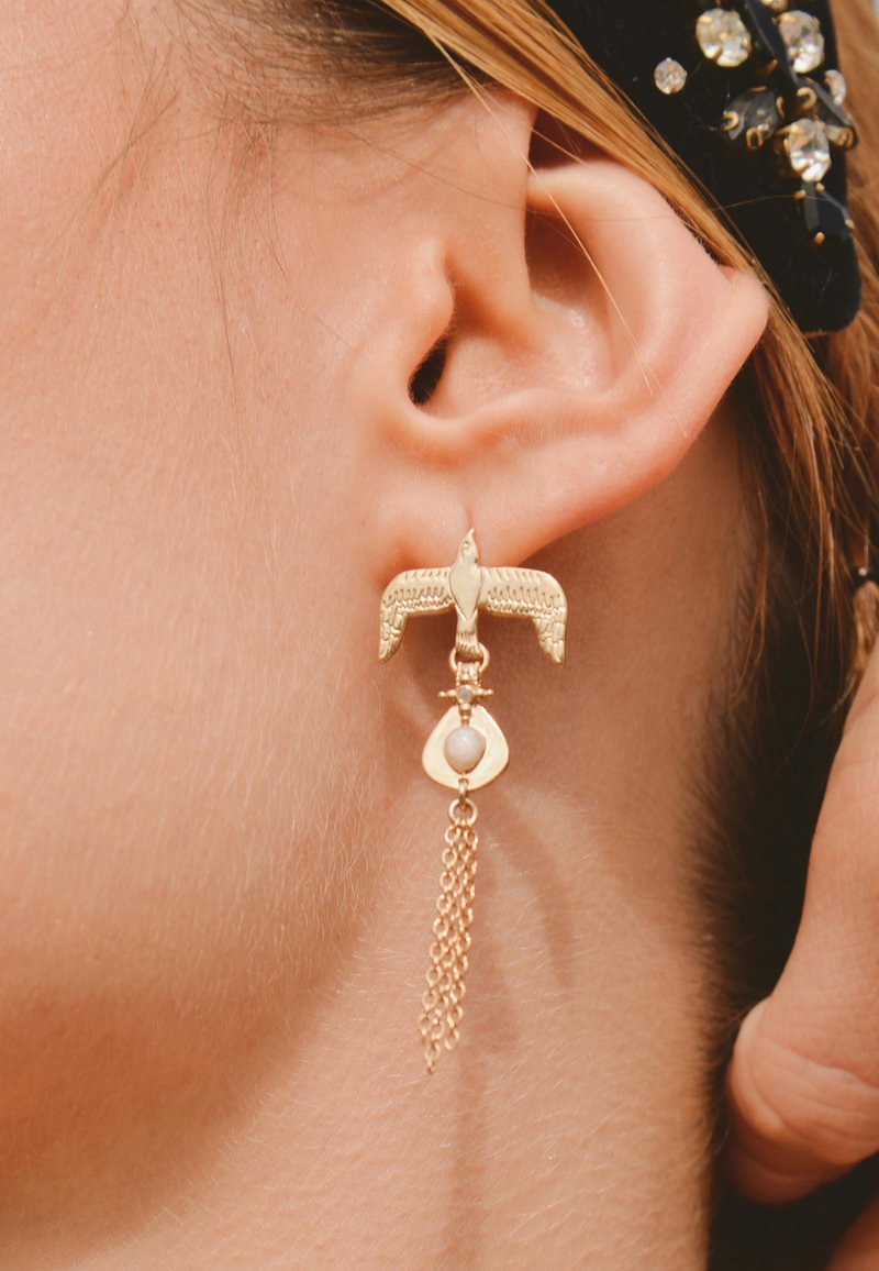 Boucles d'oreilles en or en forme d'oiseau avec un accent perlé, présentant une chaîne pendante et une goutte ouverte derrière la perle, finition texturée.