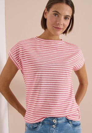 Jeune femme aux cheveux bruns courts, portant un t-shirt rayé rose et blanc et un jean bleu, debout devant un fond beige.