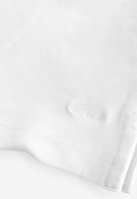Tissu en coton blanc avec une finition texturée, affichant un logo en relief subtil et des bords soigneusement cousus. Apparence lisse et légère.