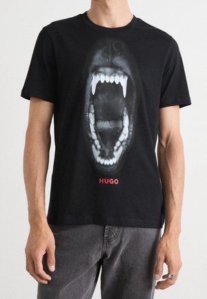 Black T-shirt with a large white graphic of an open animal mouth with sharp teeth and red "HUGO" text below, worn by a person.