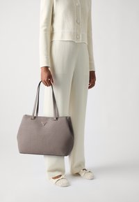 Sac fourre-tout gris texturé avec poignées doubles, présentant un détail de logo triangulaire, porté avec une veste cropped couleur crème et un pantalon ample.