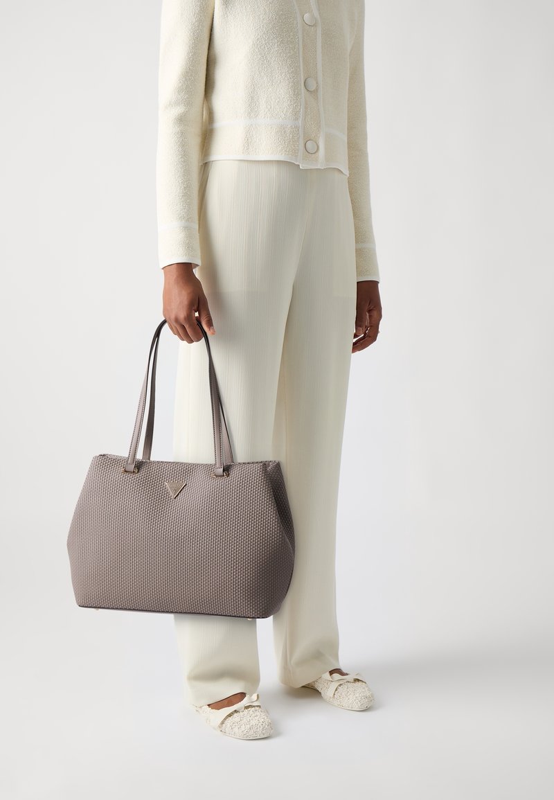 Sac fourre-tout gris texturé avec poignées doubles, présentant un détail de logo triangulaire, porté avec une veste cropped couleur crème et un pantalon ample.