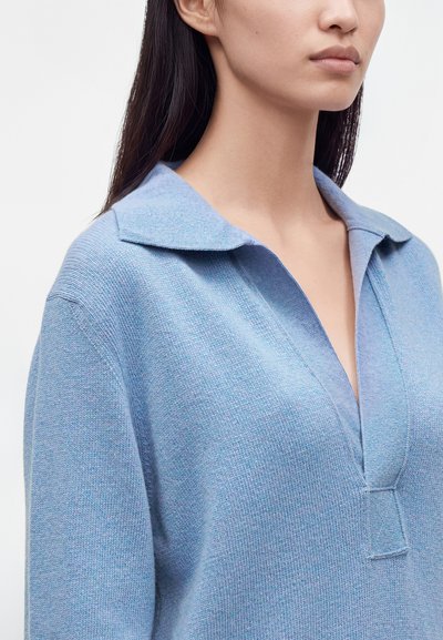 Femme portant un pull en maille bleu clair avec un large col et un décolleté en V profond, vue des épaules au menton sur un fond uni.