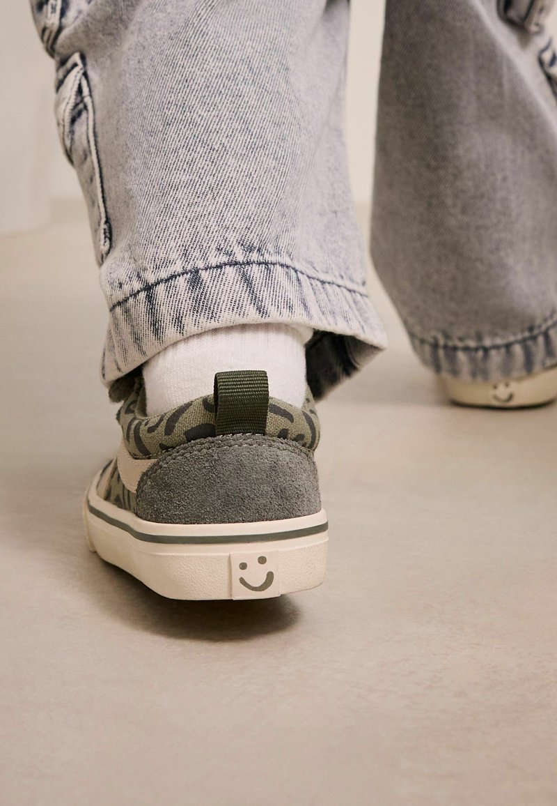 Persoon die grijze sneakers met patroon en een glimlach op de hiel draagt, loopt op een gladde vloer in licht gewassen spijkerbroek.
