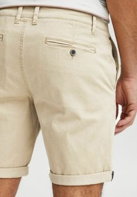 Pantalones cortos de algodón beiges con puños doblados, que cuentan con un bolsillo trasero con cierre de botón. Textura suave y ajuste entallado.