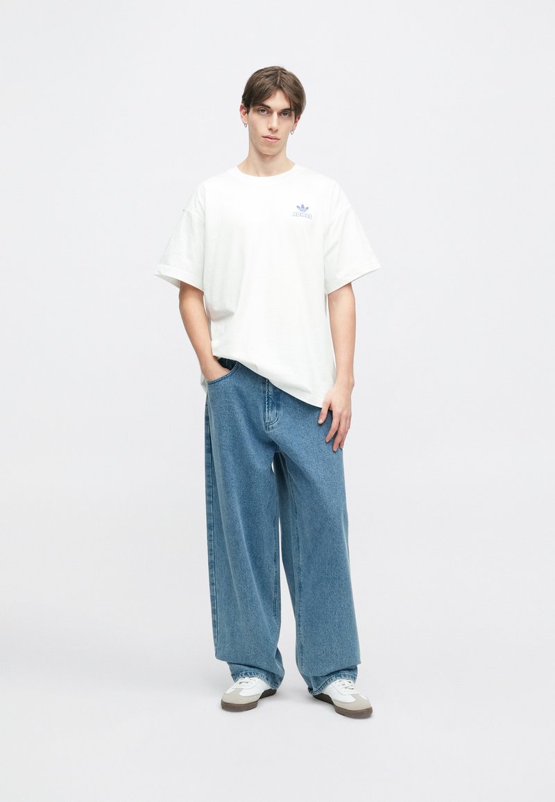 Witte oversized T-shirt met een klein blauw logo, gecombineerd met losse blauwe spijkerbroeken. De outfit omvat witte sneakers met grijze accenten.
