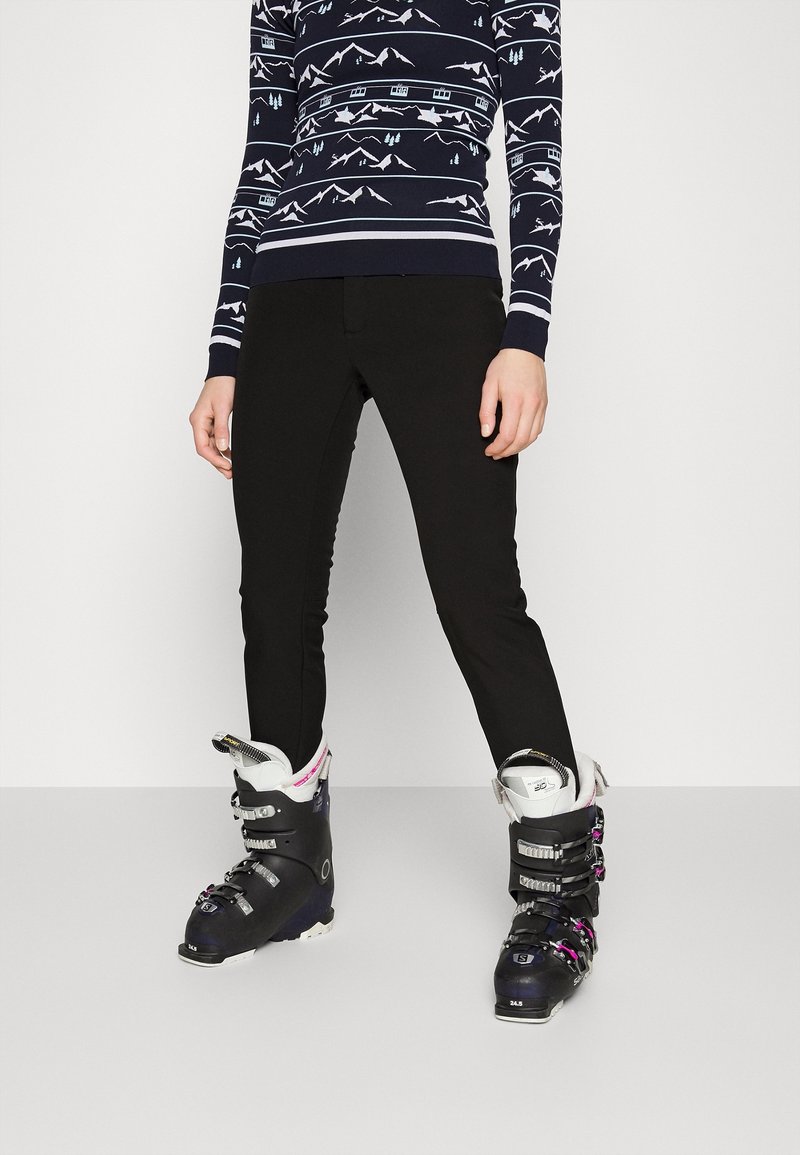 Personne portant un pantalon de ski noir, des chaussures de ski noires avec des accents blancs et roses, et un pull marine avec des motifs de montagne et d'arbres blancs.