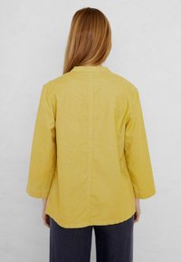 Camicia gialla leggera a maniche lunghe con colletto corto, taglio dritto e cuciture visibili. Indossata con pantaloni scuri, vista da dietro.