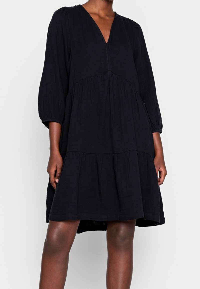 Svart lager-på-lager klänning gjord av lättviktig tyg. Har en V-ringning, ballongsärmar och en avslappnad passform. Subtil textur tillför djup.