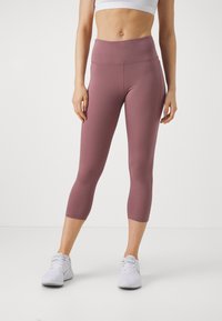 Vrouw in mauve hoge taille cropped leggings en witte sportchoenen, staande tegen een effen lichte achtergrond.