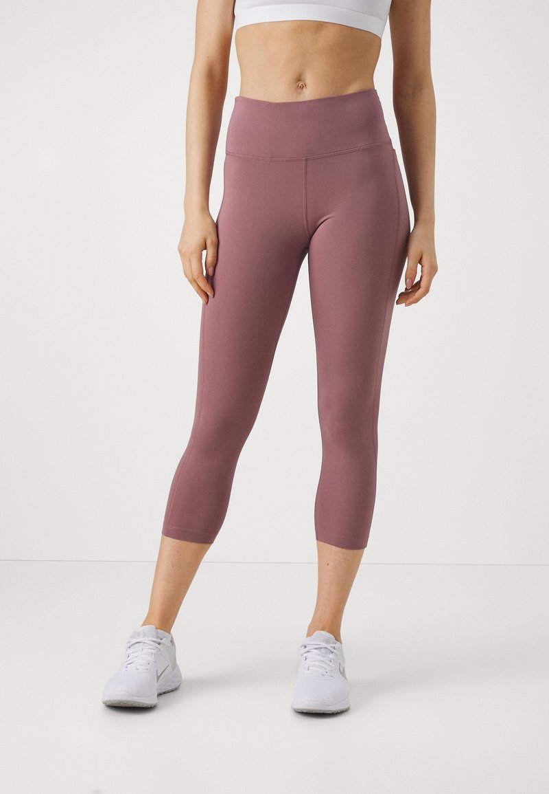 Vrouw in mauve hoge taille cropped leggings en witte sportchoenen, staande tegen een effen lichte achtergrond.