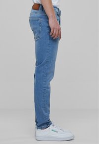 Jeans ajustados de color azul claro con una cinturilla plana, diseño de cinco bolsillos y un parche de cuero marrón en la parte trasera. Llevados con deportivas blancas.