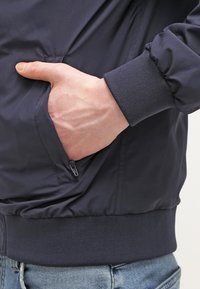 Jachetă de bomber de culoare bleumarin cu un buzunar cu fermoar pe piept, mansete tricotate și un exterior neted, lucios. Mână arătată odihnindu-se în buzunarul lateral.