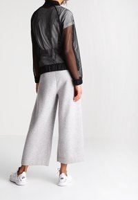 En person står med ryggen mot kameran, klädd i en svart nätjacka och grå vida byxor, tillsammans med vita sneakers.