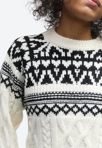 Γυναίκα που φοράει λευκό πλεκτό πουλόβερ με μαύρα γεωμετρικά και διαμαντένια σχέδια στο στήθος και στους ώμους.