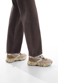 Sneakers sportivi beige con tessuto testurizzato, accenti scuri in contrasto, lacci e suola con motivo. Abbinati a pantaloni larghi marrone scuro.