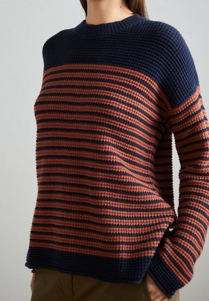 Personne portant un pull en maille texturée avec des rayures horizontales bleu marine et orange rouille, debout les mains dans les poches.