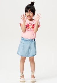 Rosa T-Shirt mit einem tropischen Print, kombiniert mit einem hellblauen Denim-Rock. Das Kind trägt beigen Espadrilles mit Ripsband-Akzenten.