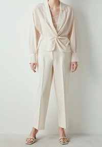 Blouse enveloppante beige clair avec des rayures texturées subtiles, associée à un pantalon plissé crème et des sandales à talons tressées avec des accents dorés.