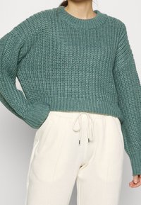 Teal stickad tröja med ribbad textur och rund halsringning, kombinerad med krämfärgade joggerbyxor som har sidfickor och dragsko i midjan.