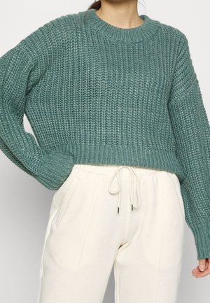 Türkis gestrickter Pullover mit Rippenstruktur und rundem Ausschnitt, kombiniert mit cremefarbenen Jogginghosen mit Seitentaschen und Kordelzugbund.