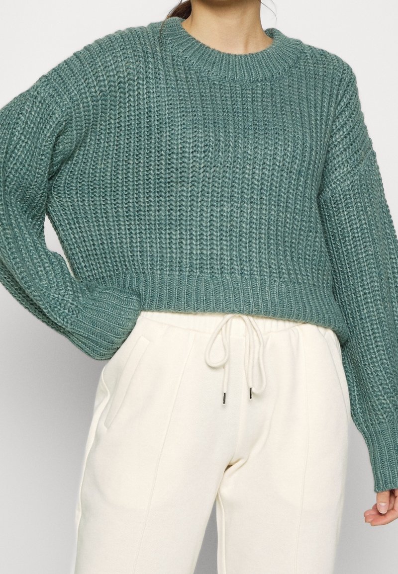 Teal stickad tröja med ribbad textur och rund halsringning, kombinerad med krämfärgade joggerbyxor som har sidfickor och dragsko i midjan.