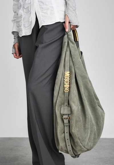 Suuri harmaa mokkahihnatasku, jossa on kultainen "MOSCHINO"-logo, säädettävä olkahihna, pehmeä tekstuuri ja rento muotoilu, yhdistettynä harmaisiin housuihin.