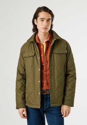 Gequiltede olijfgroene jas met drukknoopsluitingen, twee borstzakken en een puntkraag over een roestkleurig shirt. Spijkerbroeken maken de outfit compleet.
