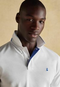 Balta polo krekls ar kontrastējošu zilu apkakli, ar pogātu plaku un mazu zilu logotipu kreisajā krūtī.