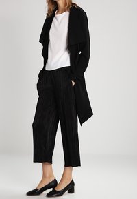 Giacca lunga nera, t-shirt bianca e culotte nere plissettate. Scarpe nere con tacco basso a blocco, texture liscia e design minimalista.