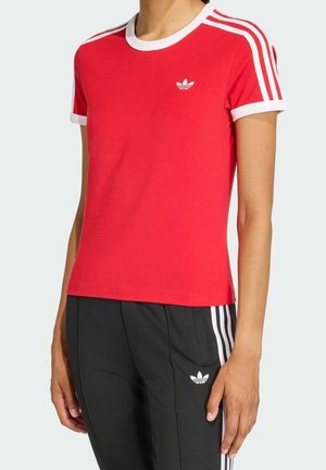 Person trägt ein rotes Adidas-T-Shirt mit weißen Streifen auf den Schultern und schwarze Adidas-Hosen mit weißen Streifen an den Seiten.