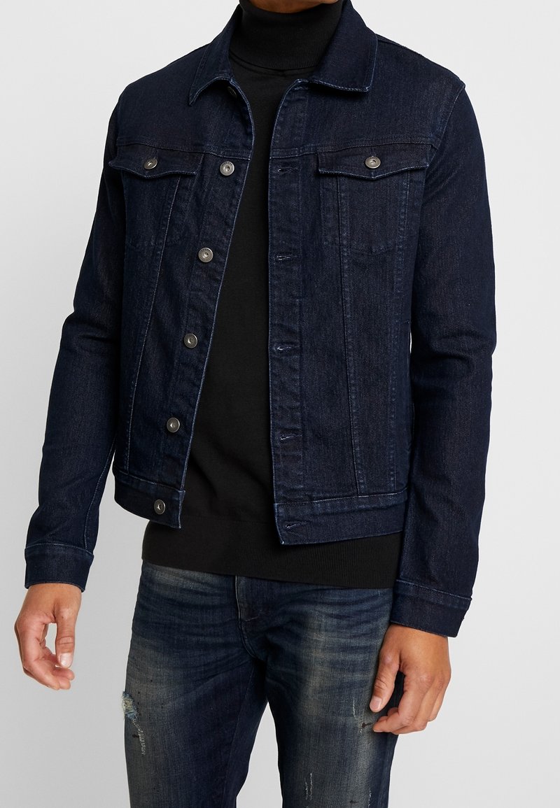 Chaqueta de mezclilla en azul oscuro con cierre de botones, dos bolsillos en el pecho y un cuello. Combinada con un suéter de cuello alto negro y jeans desgastados.