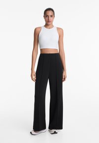Un pantalon large noir avec des plis marqués, associé à un crop top blanc à côtes. Le tissu est lisse ; des chaussures de sport complètent le look.