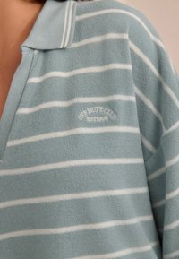 Blaustriped Polohemd mit Kragen aus weichem Stoff, mit weißen Streifen und gesticktem Logo "OFF DUTY CLUB EST.1996" auf der Brust.