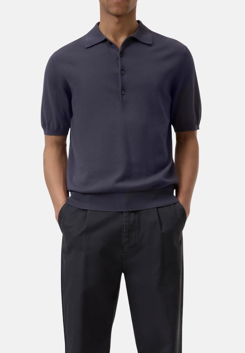 Polo a maniche corte di colore blu navy, con colletto a tre bottoni, polsini e orlo a costine; abbinato a pantaloni neri su misura per un look snello.