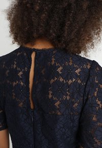 Navy lace dress showing floral pattern, short sleeves, with a back keyhole closure and small button detail at the neckline.