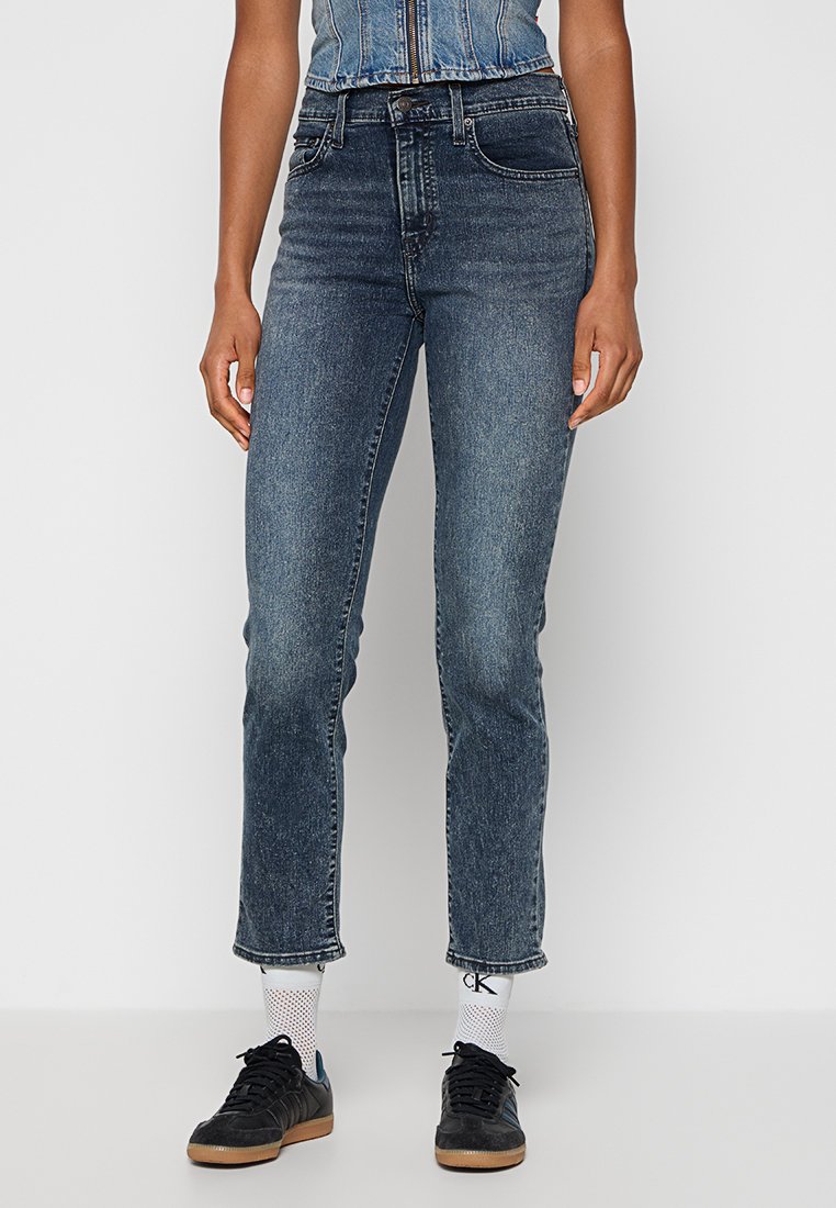 Personne portant un jean bleu taille mi-haute, des chaussettes en maille blanches avec le logo "CK", des baskets noires et un gilet en denim bleu court, se tenant devant un fond uni.