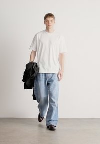 Tricou alb cu mâneci scurte, blugi largi de culoare albastru deschis și o jachetă închisă la culoare ținută într-un braț. Modelul poartă pantofi închiși la culoare și stă pe un fundal neutru.