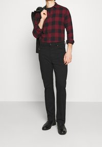 Chemise à carreaux rouge et noir à boutons, assortie à un jean noir foncé et des chaussures noires, avec une veste en cuir jetée sur une épaule.